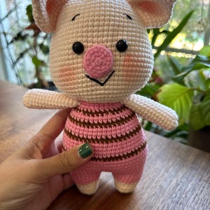 May include: A pink crocheted pig toy with brown and pink striped clothing. The pig has black eyes and a pink nose.