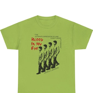 Puede incluir: Camiseta verde con un gráfico en blanco y negro de cuatro hombres caminando en fila. El texto "THE ULTIMATE EXPRESSION OF LAW IS NOT ORDER IT'S PRISON" está impreso en negro sobre el gráfico. El texto "BLOOD IN MY EYES" está impreso en rojo sobre el gráfico. El texto "GEORGE L. JACKSON" está impreso en negro debajo del gráfico.