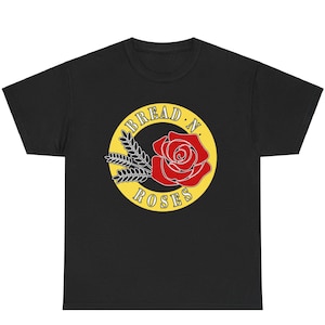 Puede incluir: Camiseta negra con un gráfico circular amarillo que presenta una rosa roja y tallos de trigo. El texto "BREAD N. ROSES" está escrito en blanco alrededor del círculo.