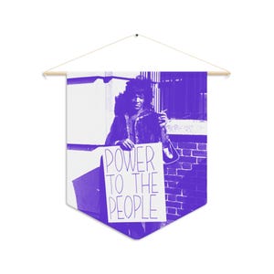Può includere: Uno stendardo da parete viola e bianco con una grafica monocromatica di una persona che tiene un cartello con la scritta POWER TO THE PEOPLE. Lo stendardo è appeso a un'asta di legno con un cordino per il fissaggio a parete.