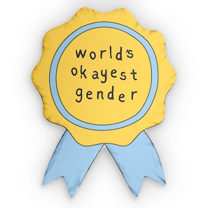 Könnte beinhalten: Ein gelber Rosetten-Abzeichen mit hellblauem Rand und Bändern. Der Text "world's okayest gender" ist in Schwarz innerhalb des blau umrandeten Kreises geschrieben. Das Abzeichen hat ein cartoonartiges Design.