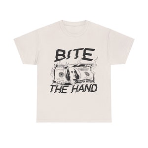 Pode incluir: Uma camiseta branca com um gráfico preto de uma nota de 100 dólares em chamas e o texto "BITE THE HAND".