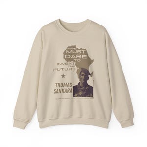Può includere: Una felpa beige con una stampa grafica marrone di un contorno dell'Africa e un ritratto di Thomas Sankara. La grafica include la citazione "Dobbiamo osare inventare il futuro." e le date "21 dicembre 1949 - 15 ottobre 1987".