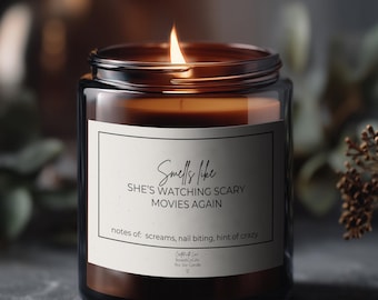 Könnte beinhalten: Eine brennende Kerze in einem dunklen bernsteinfarbenen Glasgefäß mit weißem Etikett. Der Text lautet: Smells like SHE'S WATCHING SCARY MOVIES AGAIN, notes of: screams, nail biting, hint of crazy. Die Kerze hat eine einzelne Flamme und dunkles Wachs.