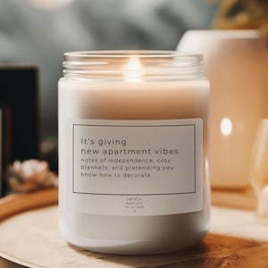 Op de afbeelding: Een brandende witte kaars in een heldere glazen pot. De kaars heeft een label met de tekst "It's giving new apartment vibes" en andere tekst. De kaars staat op een houten dienblad.