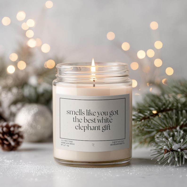 A lit candle in a glass jar with a label that reads "smells like you got the best white elephant gift" is surrounded by holiday decorations, including pine branches and ornaments, with a blurred background of lights.
