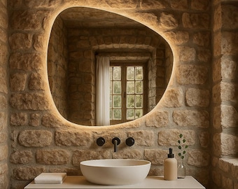 May include: A modern bathroom with a stone wall and a large, illuminated oval mirror. A white vessel sink sits atop a wooden vanity. The room includes a window, soap dispenser, and a small vase with greenery.