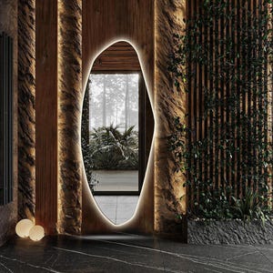 May include: A modern, irregularly shaped mirror with a wooden frame, set against a wall with a wood and stone design. The mirror reflects a view of a lush, green outdoor space.