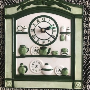 May include: A green and white ceramic wall clock with a kitchen theme. The clock features a white face with black Roman numeral hour markers and black hands. The clock is surrounded by a green frame with a white background and green accents. The background features a white shelf with green teapots, cups, and saucers.