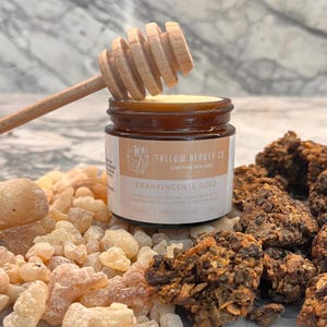 May include: A jar of Frankincense Gold balm with a wooden honey dipper. The jar is surrounded by pieces of frankincense resin and other natural ingredients. The label reads "Tallow Beauty Co" and "Low Pufa Skin Care."