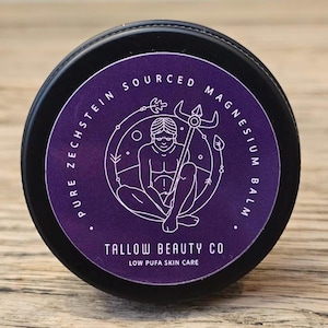 Puede incluir: Un recipiente negro redondo con una etiqueta morada que presenta texto blanco que dice Pure Zechstein Sourced Magnesium Balm y Tallow Beauty Co Low Pufa Skin Care. El gr&aacute;fico central muestra una ilustraci&oacute;n lineal de una figura sentada con un bast&oacute;n.