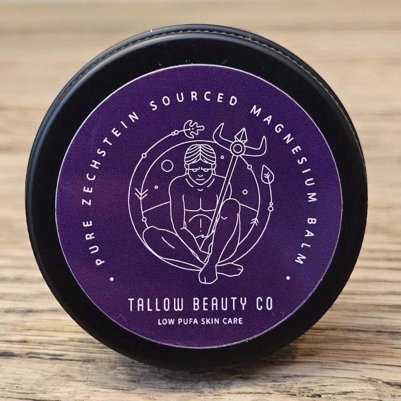 Puede incluir: Un recipiente negro redondo con una etiqueta morada que presenta texto blanco que dice Pure Zechstein Sourced Magnesium Balm y Tallow Beauty Co Low Pufa Skin Care. El gr&aacute;fico central muestra una ilustraci&oacute;n lineal de una figura sentada con un bast&oacute;n.