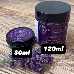 Puede incluir: Dos tarros negros de b&aacute;lsamo de magnesio Sacred Sleep de Tallow Beauty Co., uno de 30ml y otro de 120ml, con etiquetas moradas, texto blanco, una ilustraci&oacute;n de l&iacute;neas y una rama de flores de lavanda moradas frescas.