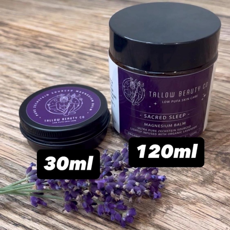 Puede incluir: Dos tarros negros de b&aacute;lsamo de magnesio Sacred Sleep de Tallow Beauty Co., uno de 30ml y otro de 120ml, con etiquetas moradas, texto blanco, una ilustraci&oacute;n de l&iacute;neas y una rama de flores de lavanda moradas frescas.