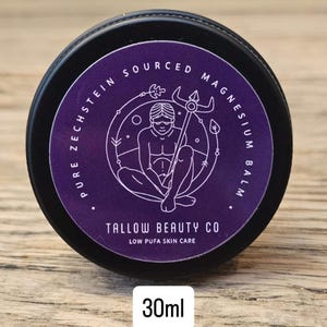 Puede incluir: Un recipiente negro redondo de b&aacute;lsamo de magnesio Tallow Beauty Co. con una etiqueta morada. La etiqueta presenta un dibujo lineal blanco de una figura sosteniendo un tridente, rodeado por el texto Pure Zechstein Sourced Magnesium Balm, 30ml.
