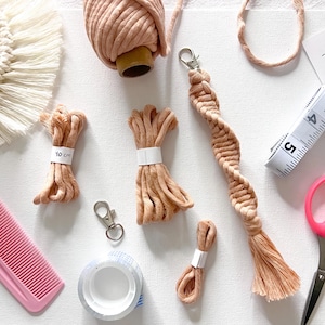 Puede incluir: Una colección de suministros para macramé, que incluye cordón beige, un peine rosa, tijeras, una cinta métrica y un llavero. Los paquetes de cordón están etiquetados con cinta blanca. También se ve una nota manuscrita.