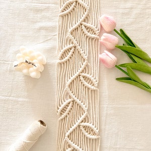 Puede incluir: Colgante de pared de macramé color crema con diseño de hojas, montado en una varilla de madera. Se muestra con tulipanes rosas y una vela blanca. También se ve un carrete de hilo color crema.