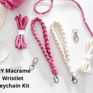 May include: A DIY macrame wristlet keychain kit with pink and cream-colored materials. The kit includes macrame cord, finished wristlets, and silver-colored lobster clasps. The text "DIY Macrame Wristlet Keychain Kit" is visible.