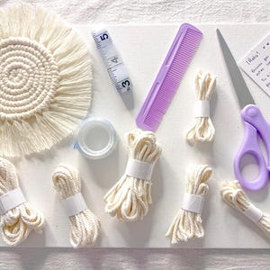 Puede incluir: Un kit de manualidades de macramé con un posavasos color crema, paquetes de cuerda color crema, una cinta métrica, un peine morado, tijeras y cinta adhesiva. También se incluye una nota manuscrita. El kit está dispuesto sobre una superficie blanca.