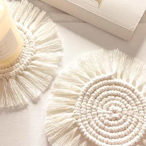 Puede incluir: Dos posavasos de macramé blanquecino, uno con una vela. Los posavasos tienen un diseño circular con un borde con flecos. Una vela en un tarro de cristal se encuentra sobre un posavasos. La etiqueta de la vela dice "SOOTHE" y "APAISE".