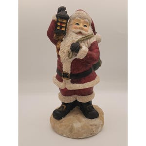 May include: A ceramic Santa Claus figurine, dressed in a red suit with white faux fur trim. He holds a black lantern in one hand and a sack over his shoulder. The figurine stands on a textured, beige base, perfect for holiday decor.