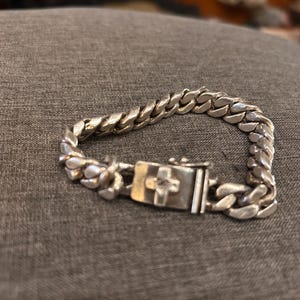 Puede incluir: Una pulsera de cadena de plata con un cierre en forma de cruz. La pulsera está hecha de eslabones entrelazados y tiene un acabado pulido. El cierre presenta un diseño de cruz.