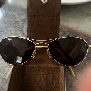 May include: Gold-rimmed sunglasses with dark lenses rest in an open brown leather case. The case is embossed with "OLIVER PEOPLES" and secured with a silver snap. The sunglasses have a classic aviator style.