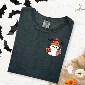Pu&ograve; includere: Una maglietta grigia scura con un simpatico cartone animato di un fantasma che indossa un cappello di lana e tiene una zucca e una tazza. Il fantasma indossa un cappello rosso e arancione e ha un'espressione amichevole. Il testo "COMFORT COLORS" e "XL - TG - EG" &egrave; stampato su un'etichetta bianca attaccata alla maglietta.