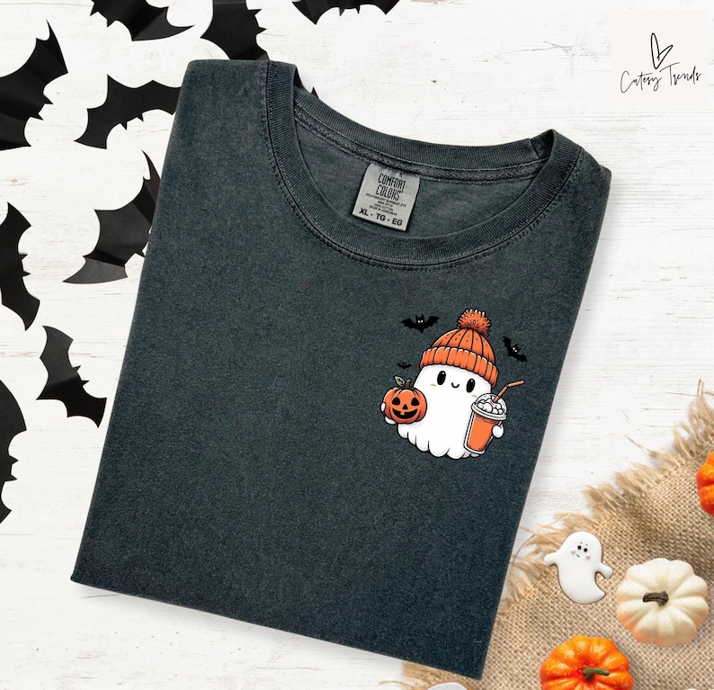 Pu&ograve; includere: Una maglietta grigia scura con un simpatico cartone animato di un fantasma che indossa un cappello di lana e tiene una zucca e una tazza. Il fantasma indossa un cappello rosso e arancione e ha un'espressione amichevole. Il testo "COMFORT COLORS" e "XL - TG - EG" &egrave; stampato su un'etichetta bianca attaccata alla maglietta.