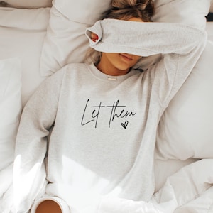 以下が含まれることがあります： 黒い文字で「Let them」と書かれたグレーのトレーナー。小さなハートのマークが付いている。