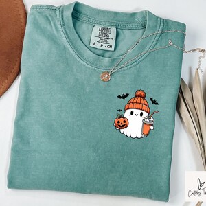 Pu&ograve; includere: Una maglietta blu-verde con un simpatico fantasma dei cartoni animati che tiene una zucca e una tazza di cioccolata calda. Il fantasma indossa un cappello rosso e bianco. La maglietta ha il testo "Comfort Colors" e "S - P - CH" stampato sull'etichetta.