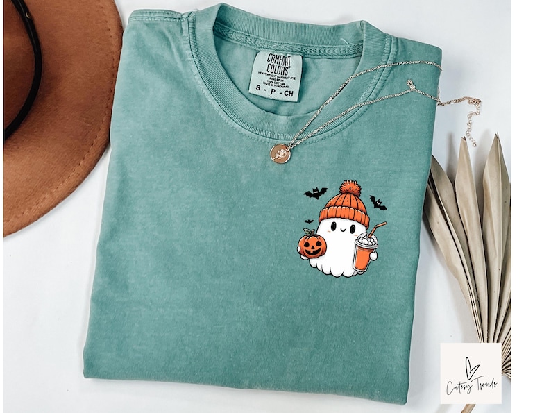 Pu&ograve; includere: Una maglietta blu-verde con un simpatico fantasma dei cartoni animati che tiene una zucca e una tazza di cioccolata calda. Il fantasma indossa un cappello rosso e bianco. La maglietta ha il testo "Comfort Colors" e "S - P - CH" stampato sull'etichetta.