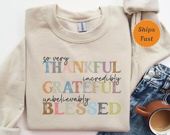 May include: A beige sweatshirt with the text "so very THANKFUL incredibly GRATEFUL unbelievably BLESSED" in colourful letters. The letters have a distressed, vintage look.