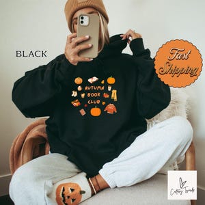 Op de afbeelding: Zwart hoodie met een grafisch ontwerp van herfstthema illustraties en de tekst "Autumn Book Club".