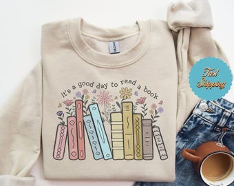 Può includere: Una felpa beige con la scritta "it's a good day to read a book" stampata sul davanti. Il testo è circondato da una pila di libri colorati e illustrazioni floreali.