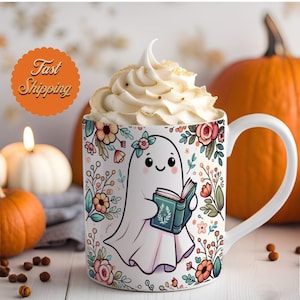 Op de afbeelding: Een witte keramische mok met een bloemenpatroon en een cartoon-spook dat een boek vasthoudt. Het spook heeft een glimlachend gezicht en draagt een wit laken. De mok is gevuld met een witte slagroomtopping.