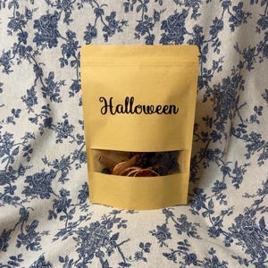 May include: A tan kraft paper stand-up pouch with a clear rectangular window at the bottom. The word Halloween is printed in black script on the front. The pouch contains dried orange slices, cinnamon sticks, and star anise pieces.