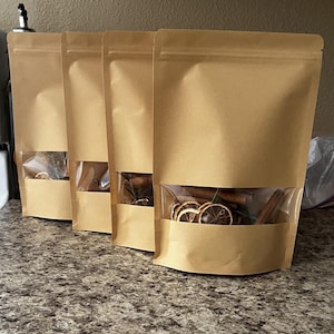 May include: Four brown paper pouches with clear windows, each filled with dried orange slices, cinnamon sticks, and other spices. The pouches are sealed and arranged in a row on a speckled countertop.