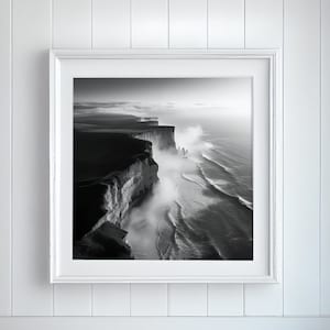 Puede incluir: Una fotografía en blanco y negro de un acantilado rocoso con olas rompiendo contra la costa. La foto está enmarcada en un marco blanco y colgada en una pared blanca.