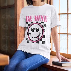 May include: White t-shirt with a black and white checkered pattern and a pink smiley face with hearts for eyes. The text "BE MINE" is printed in black above the smiley face.