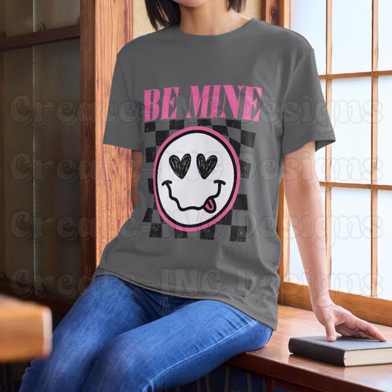 May include: A gray t-shirt with a black and white checkered pattern and a pink smiley face with hearts for eyes. The text "BE MINE" is printed in pink above the smiley face.