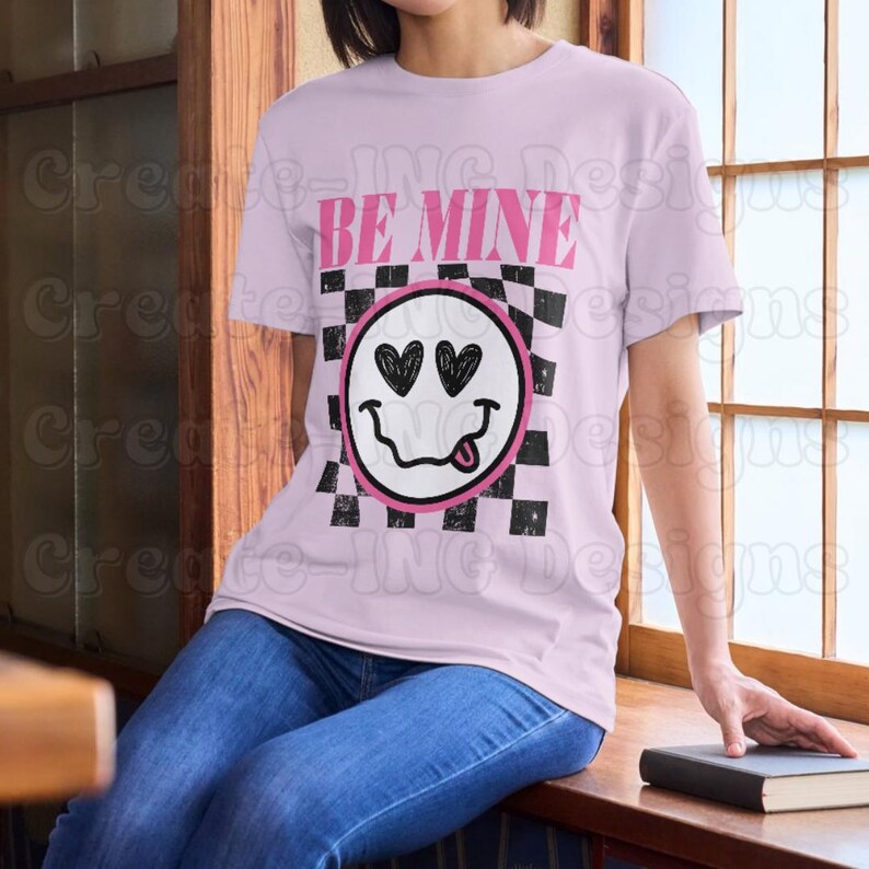 May include: Pink t-shirt with a black and white checkered pattern and a smiley face with hearts for eyes. The text "BE MINE" is printed above the smiley face.
