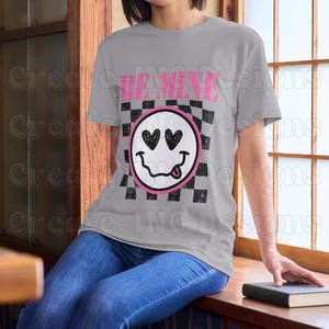 May include: A gray t-shirt with a black and white checkered pattern and a pink smiley face with hearts for eyes. The text "BE MINE" is printed above the smiley face.