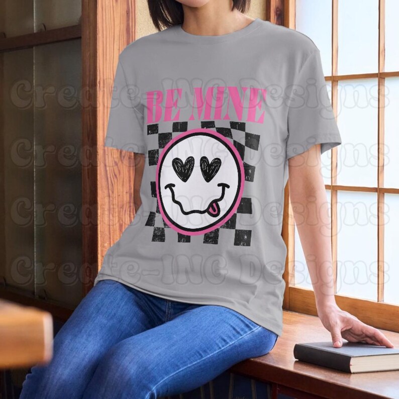 May include: A gray t-shirt with a black and white checkered pattern and a pink smiley face with hearts for eyes. The text "BE MINE" is printed above the smiley face.