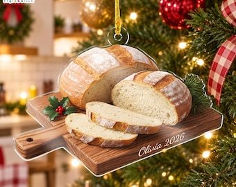 May include: A clear acrylic Christmas ornament shaped like a wooden cutting board with a loaf of bread and two slices on top. The text Olivia 2026 is printed on the board. It features a gold hanging string and is displayed on a tree.