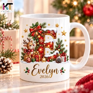May include: A white ceramic mug featuring a large red letter E decorated with snow, a red bow, and pine branches. A cartoon reindeer wearing a scarf stands next to wrapped gifts. The name Evelyn and the year 2026 are printed in gold script.