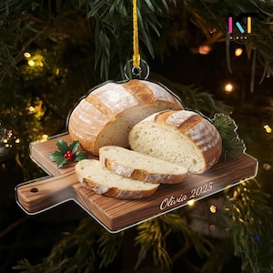 May include: A bread-themed Christmas ornament featuring a loaf of bread, a sliced loaf, and several slices on a wooden cutting board. The ornament includes the text "Olivia 2025" and is suspended by a gold string.