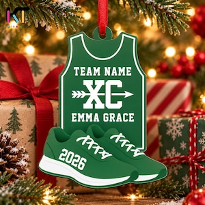 May include: A green, jersey-shaped Christmas ornament featuring the white text TEAM NAME, XC with an arrow graphic, and EMMA GRACE. Below, a pair of green running shoes with white laces and soles displays the white year 2026 on the side.