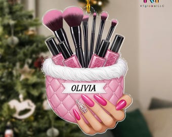 May include: A pink Christmas ornament featuring makeup brushes, nail polish, and a hand with long, pink nails. The ornament has the name "OLIVIA" on it. The background is a blurred Christmas tree.
