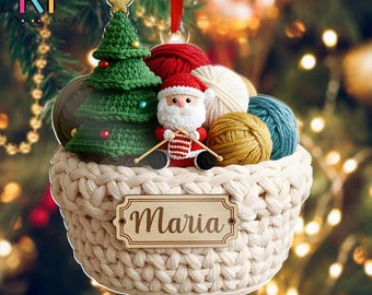 May include: A handmade Christmas ornament featuring a crocheted basket filled with yarn balls in various colors. A crocheted Santa Claus figure is knitting, and a green Christmas tree sits beside him. The ornament has a wooden name tag that says "Maria".